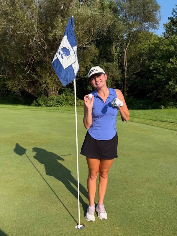 Alivia on a golf green, smiling and holding up her golf ball next to the flagstick and the cup
