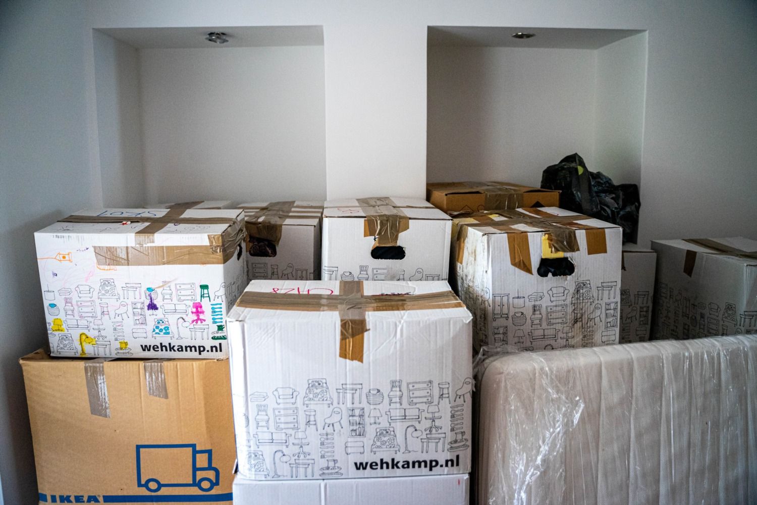 Taped moving boxes stacked against a white wall next to a mattress wrapped in plastic