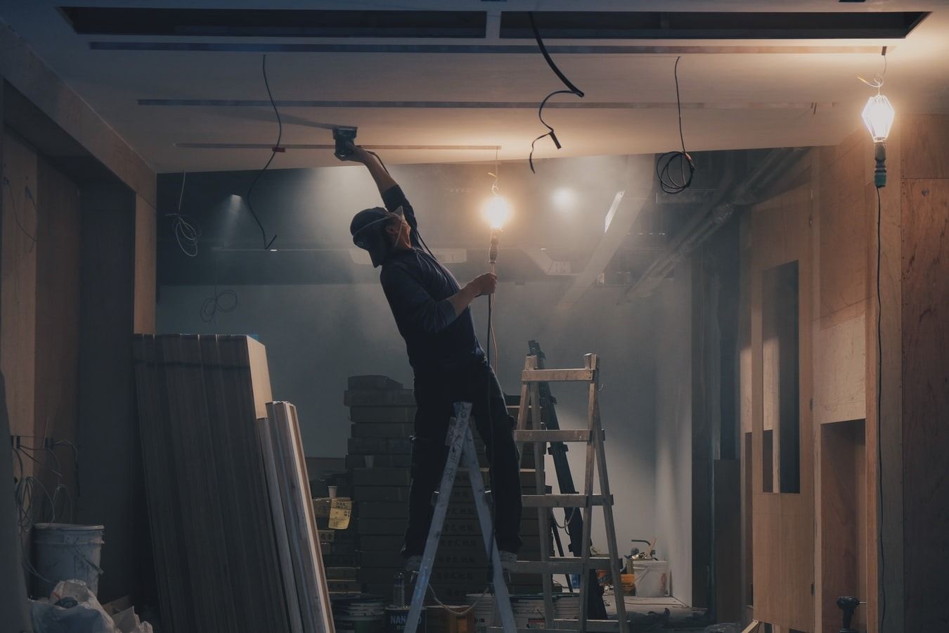 A worker on a stepladder sanding the ceiling of a gutted room under bare work lights
